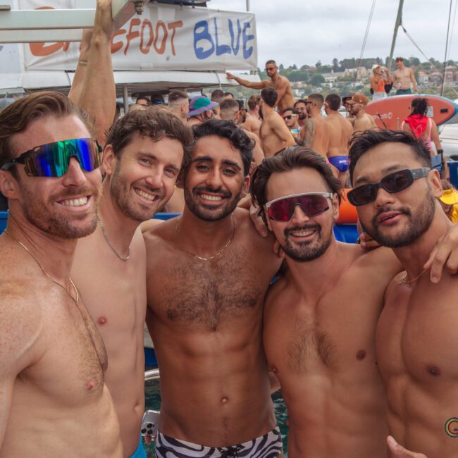 Five shirtless men are smiling and posing together on a boat at a lively outdoor party. Other people enjoy the event in the background, with boats and water visible. The atmosphere is festive and energetic.