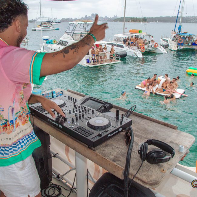 A DJ in a colorful shirt plays music on a yacht, while people swim and relax on floats and other boats nearby. The atmosphere is lively with multiple boats and partygoers enjoying the water.