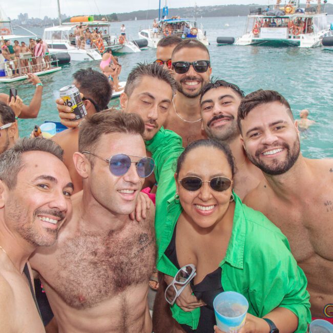A group of smiling friends pose for a photo at a lively boat party, surrounded by other people and boats on the water. Many are shirtless and holding drinks, enjoying the festive atmosphere.