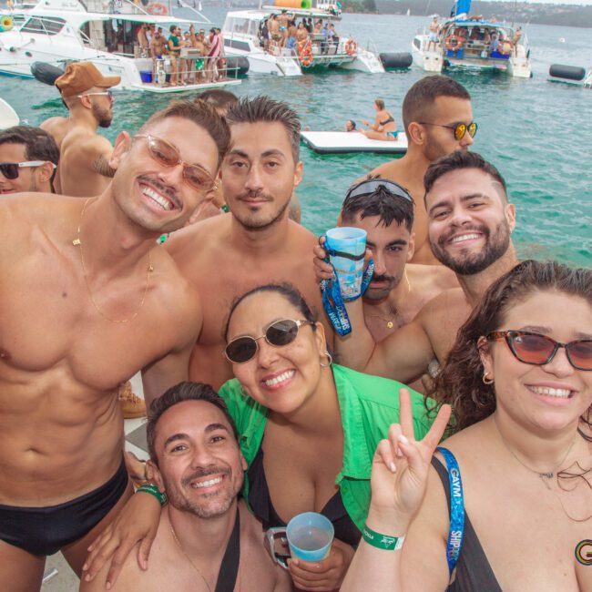A group of people in swimsuits smile and pose for a photo on a boat, with other boats and people celebrating on the water in the background. The mood is festive and sunny.