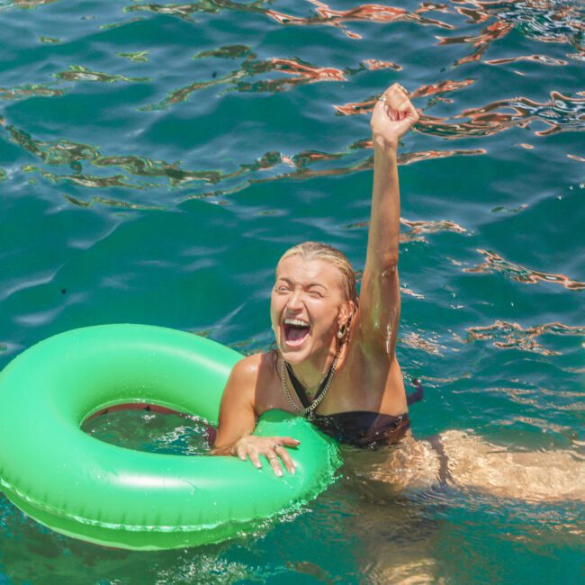 A woman in a black swimsuit floats in bright blue water with a green inflatable ring, smiling and raising one arm in excitement. Sunlight sparkles on the water's surface.