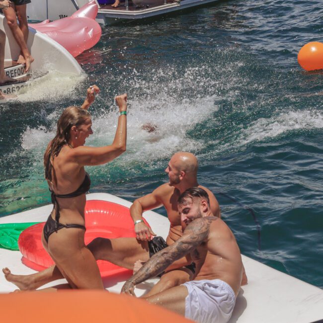 Four people sit and play on a floating mat in the water near boats, with one woman in a black bikini raising her arms. An orange float and splash are visible in the background. The atmosphere is lively and fun.