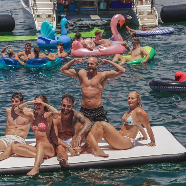 A group of people in swimsuits pose and smile on a floating mat in the water, surrounded by others relaxing on colorful inflatable pool floats near boats on a sunny day.