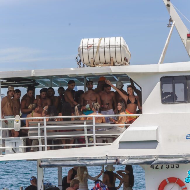 A group of people in swimsuits enjoy a party on the deck of a white boat called "Barefoot," with one woman looking out from a window. City buildings and palm trees are visible in the background under a clear sky.