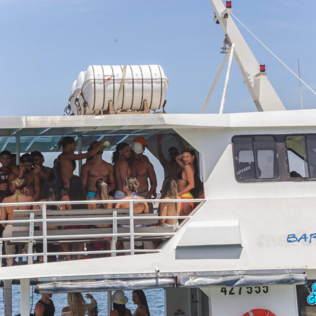 A group of people in swimsuits are standing and sitting on the deck of a white boat named "Barefootcat," enjoying a sunny day on the water. Some are chatting while others look out at the view.