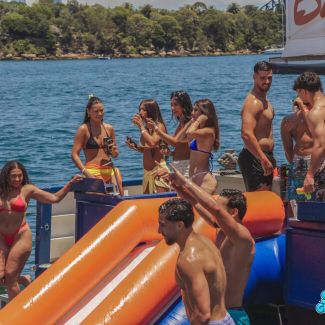 A group of people in swimsuits enjoy a sunny day on a boat, some standing, talking, or holding drinks, while others are near an inflatable slide over the water. There are trees and city buildings in the background.