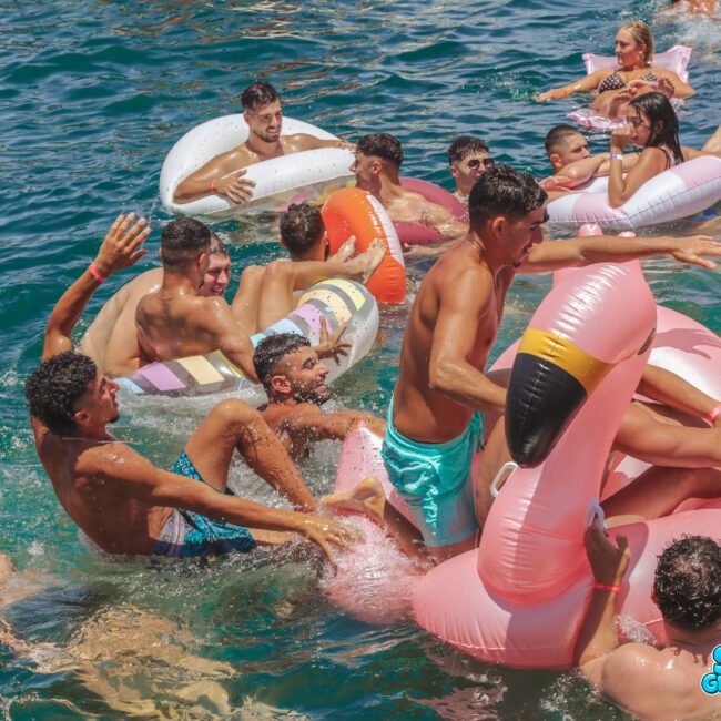 A group of young people in swimsuits laugh and play in the water with colorful inflatable floats, including a large pink flamingo, as sunlight reflects off the blue water. The mood is joyful and energetic.