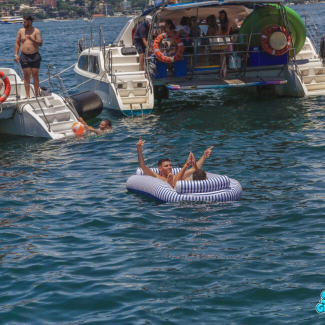 Two people are relaxing on an inflatable mattress in the water near two docked boats filled with people enjoying a sunny day. The atmosphere is festive, with blue water and clear skies.