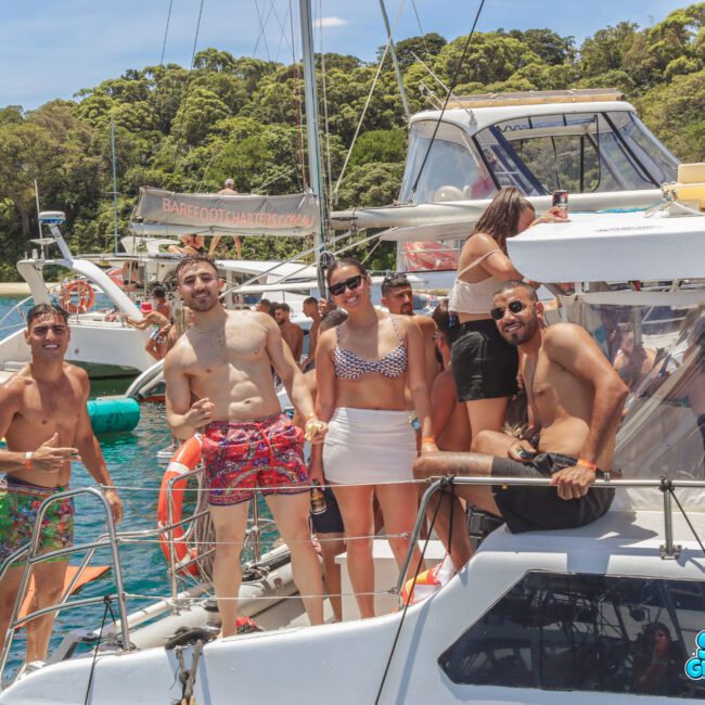 A group of five people in swimwear smile and pose on a boat, surrounded by water and other boats. Lush green trees are visible in the background under a sunny sky.