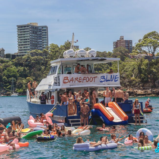 A large group of people in swimsuits enjoy a sunny day on a boat and in the water with inflatables. The boat has a "Barefoot Blue" banner, and tall buildings and trees are visible in the background.