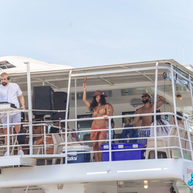 A group of people dance and DJ on the upper deck of a white boat under a clear sky. Some wear swimsuits and hats, and there is music equipment and coolers visible. The logo "Sail Gustavo" is on the lower right.