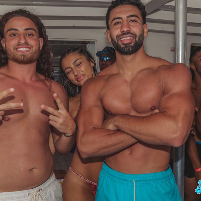 A group of young adults in swimwear smiling and posing for a photo on a boat, with two men in front, one flashing a peace sign. The image has "Sail Gustavo" and "Lush Visual" watermarks.