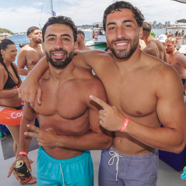 Two men in swim trunks smile and pose for the camera on a boat, one with his arm around the other. People in swimsuits and boats are in the background, suggesting a lively party atmosphere on the water.