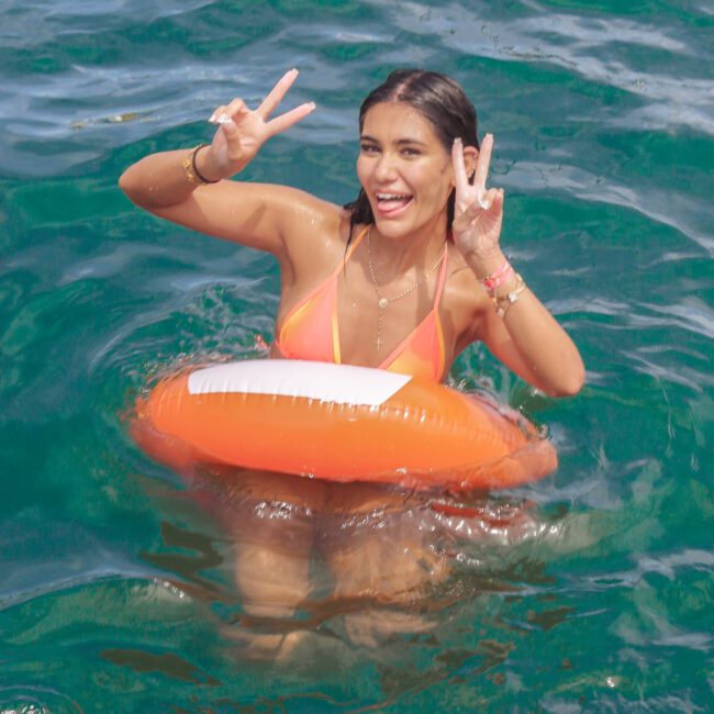 A woman in an orange swimsuit and inflatable ring smiles and makes peace signs with both hands while standing in clear blue water near a dock. The logo “Sail Gustavo” is visible in the bottom right corner.