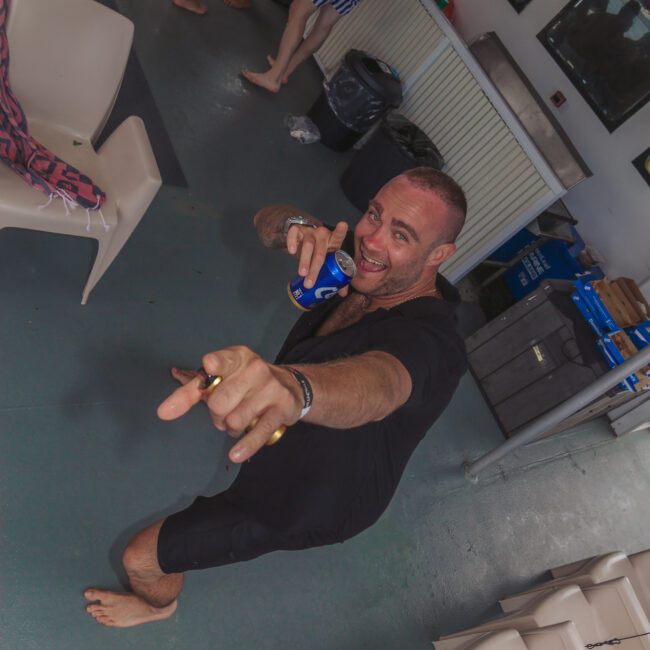 A barefoot man in a black shirt enthusiastically points at the camera while holding a drink on a boat. Other people in striped swim trunks stand and socialize in the background. The scene appears lively and fun.