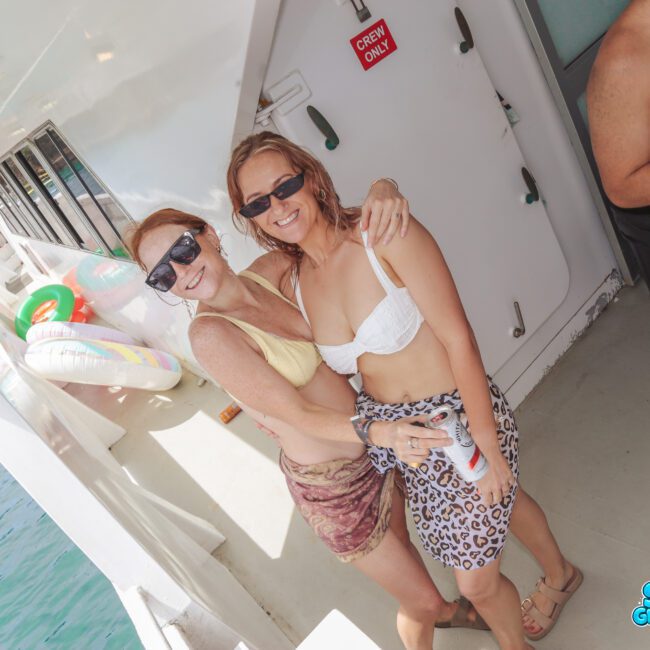 Two women in swimsuits and sunglasses smile and hug on a boat deck. Another person stands nearby holding a drink. Pool floats and clear blue water are visible in the background. The sun is shining brightly.