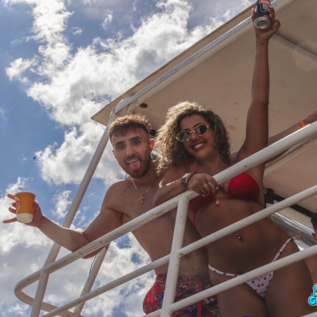 Two people in swimsuits stand on a boat deck, smiling and posing with drinks in their hands under a partly cloudy sky. The woman wears sunglasses and a red bikini. "Sail Gustavo" logo appears in the bottom right corner.