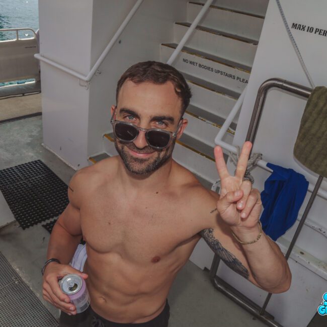 A smiling, shirtless man with sunglasses holds a drink and flashes a peace sign aboard a boat, with towels and stairs in the background. The water and boat railing are also visible.