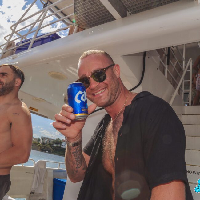 A man in sunglasses and an open shirt smiles while holding a can of beer on a boat. Other people in swimwear are around him, and the sky is bright and sunny. The boat stairs and water are visible in the background.
