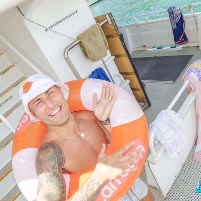 A smiling man with a white hat and tattoos poses on a boat while wearing an orange inflatable swim ring. Towels hang nearby, and water can be seen in the background. The logo "Sail Gustavo" is in the bottom right corner.
