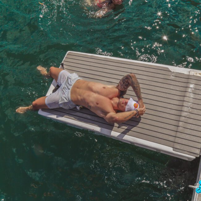 A man in swim trunks lies on his back on a dock over green water, covering his face with his arm. Another swimmer is visible nearby in the water. The photo has a watermark that says "Sail Gustavo.