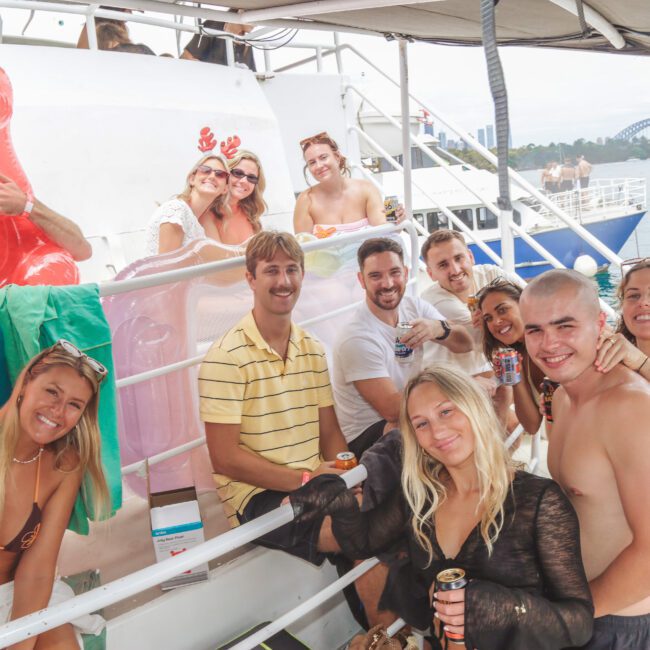 A group of young adults smile and pose together on a boat, holding drinks. They are casually dressed for warm weather, with pool floats and towels visible. The water and a bridge can be seen in the background.