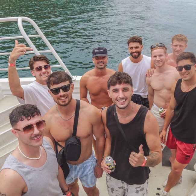 A group of nine young men, mostly shirtless and wearing sunglasses, smile and pose together on a boat with water in the background. Some hold drinks, and the weather appears sunny and warm.