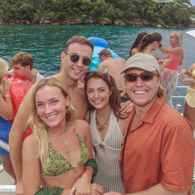 A group of smiling young adults pose for a photo on a boat. They are wearing summer clothes and sunglasses, with water and trees in the background. Other people are mingling and relaxing behind them.