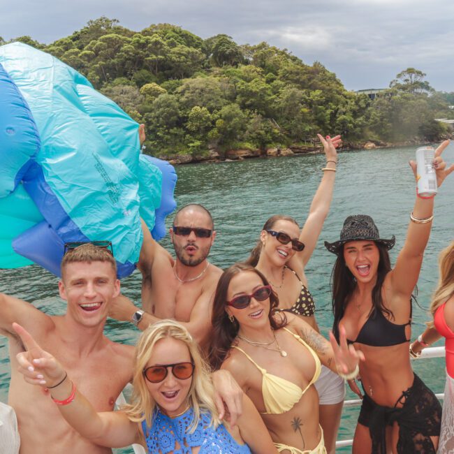 A group of young adults in swimwear pose and smile on a boat, holding drinks and making peace signs, with trees and water in the background and a large blue inflatable in the corner.
