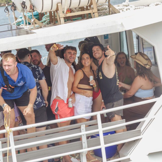 A group of people enjoy a lively party on a boat, smiling, posing, and holding drinks. Some wear swimsuits and sunglasses, while others are dressed casually. The atmosphere is festive and relaxed near the water.