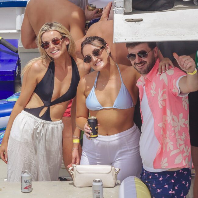 Three people in swimsuits and sunglasses smile at the camera on a boat, holding drinks. The scene is sunny and festive, with other guests and floatation devices visible in the background.