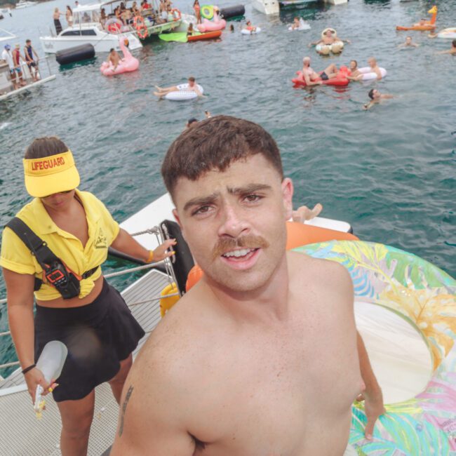 A shirtless man stands on a dock next to a colorful inflatable ring. A lifeguard in a yellow shirt and hat stands nearby. Dozens of people on inflatables and boats enjoy a lively day on the water in the background.