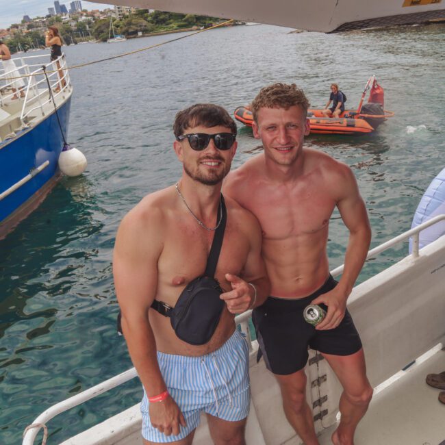 Two men in swimwear stand smiling on a boat, one wearing sunglasses and a crossbody bag, the other holding a drink. Water and another boat are visible in the background, with people on an inflatable and a dinghy.