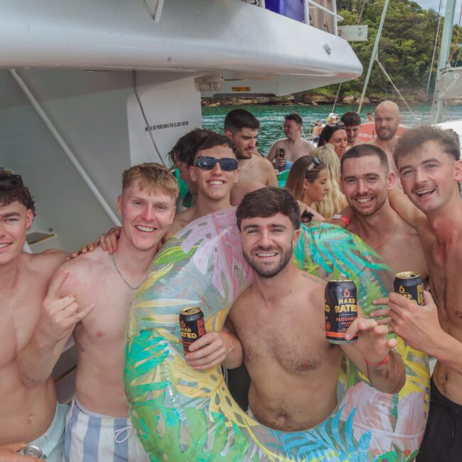 A group of young men in swimwear pose together on a boat, smiling and holding cans of beer. One has an inflatable pool float around him. Other people and greenery are visible in the background.