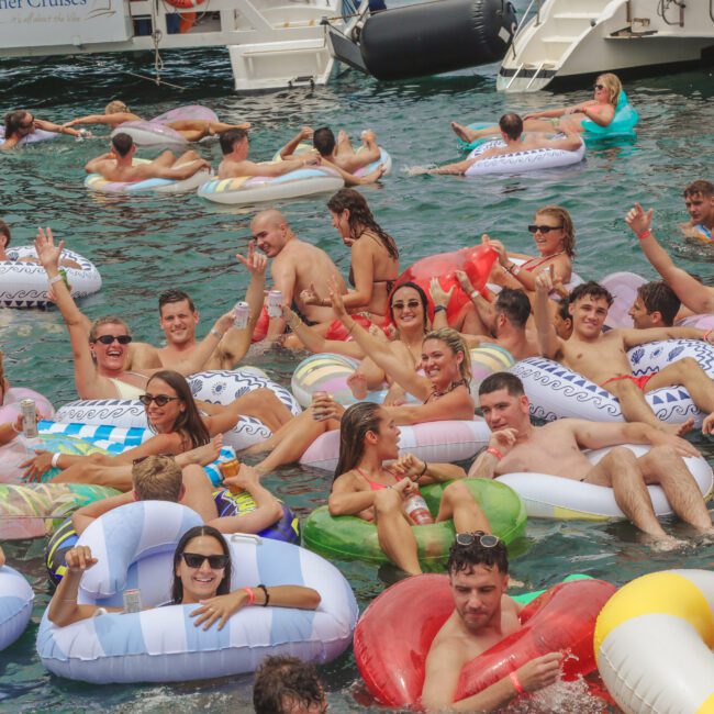 A large group of people on colorful inflatable floats enjoy a lively party in the water near a docked boat, smiling, waving, and holding drinks. The atmosphere is festive and relaxed.
