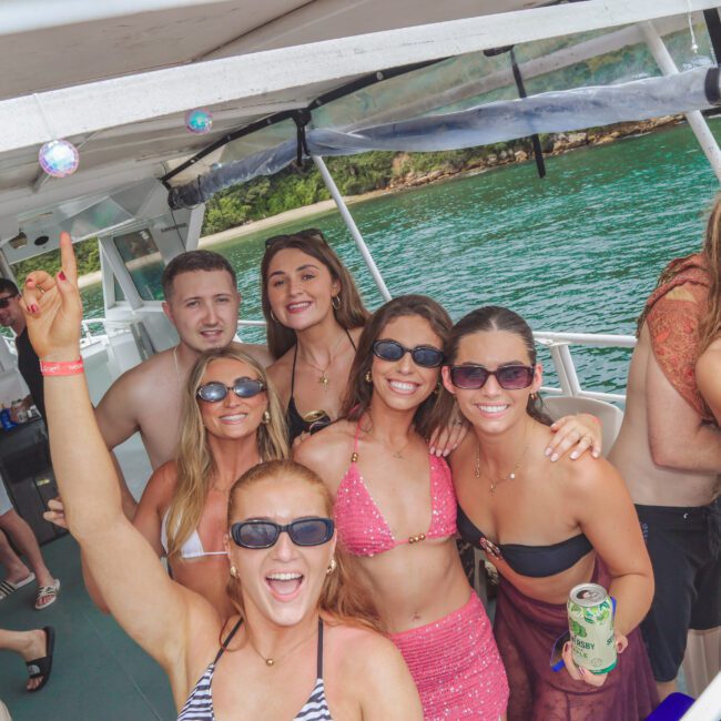 A group of smiling people in swimwear and sunglasses pose on a boat, enjoying a sunny day on the water. Some hold drinks, and greenery is visible in the background.