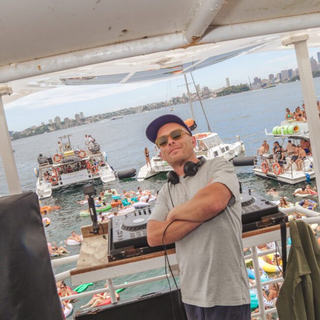 A DJ in sunglasses and a cap stands at a mixing console on a boat, arms crossed, with a lively crowd of people on inflatables and boats enjoying a party on the water in the background.
