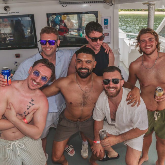 Six smiling men in swimwear pose together on a boat, holding drinks. The boat's interior and windows showing water and greenery are visible in the background. The mood is casual and festive.