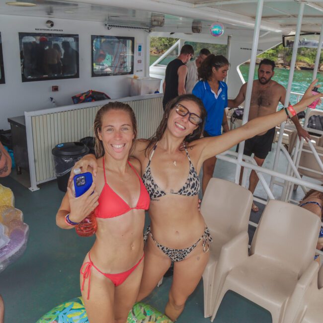 Two women in bikinis smile and pose for the camera on a boat, surrounded by other people enjoying a party near the water. The atmosphere is lively, with sunlight, swimsuits, and inflatable floats visible.