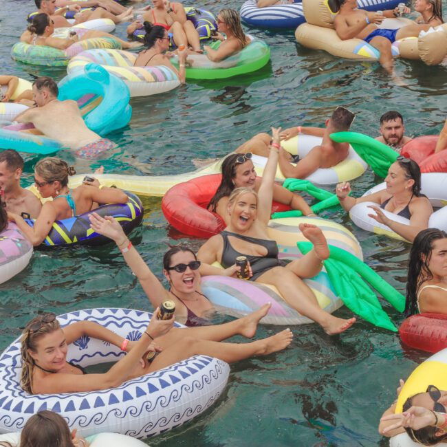 A large group of people relax on colorful inflatable floats in a pool, smiling, holding drinks, and enjoying the sun. The scene is lively and festive, with various float shapes and many people interacting.