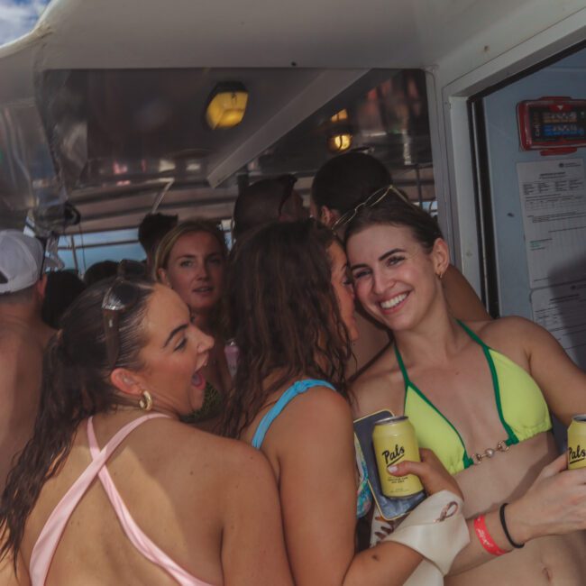 A group of young women in swimsuits smile and pose together on a boat, holding drinks, with other people and a blue sky visible in the background.