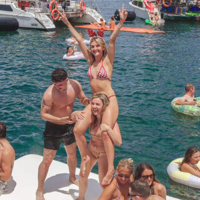 A group of people in swimsuits enjoy a lively boat party on the water. One woman sits on another’s shoulders with arms raised, surrounded by others relaxing on floaties and boats in the background.