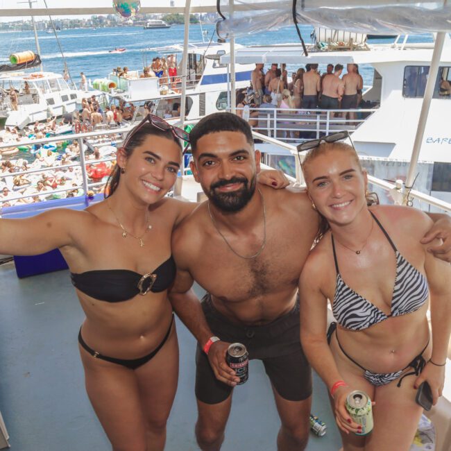Three people in swimsuits smile and pose with drinks on the deck of a boat, with a large crowd, more boats, and water in the background on a sunny day.