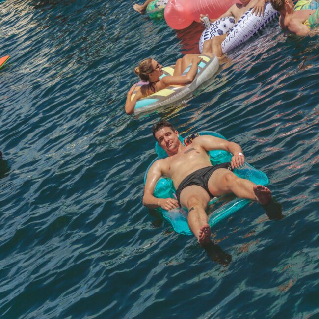 A man relaxes on a blue inflatable float in the water, surrounded by other people on colorful pool floats. The sun is shining and the atmosphere is festive. The "Yacht Social Club" logo is in the bottom right corner.