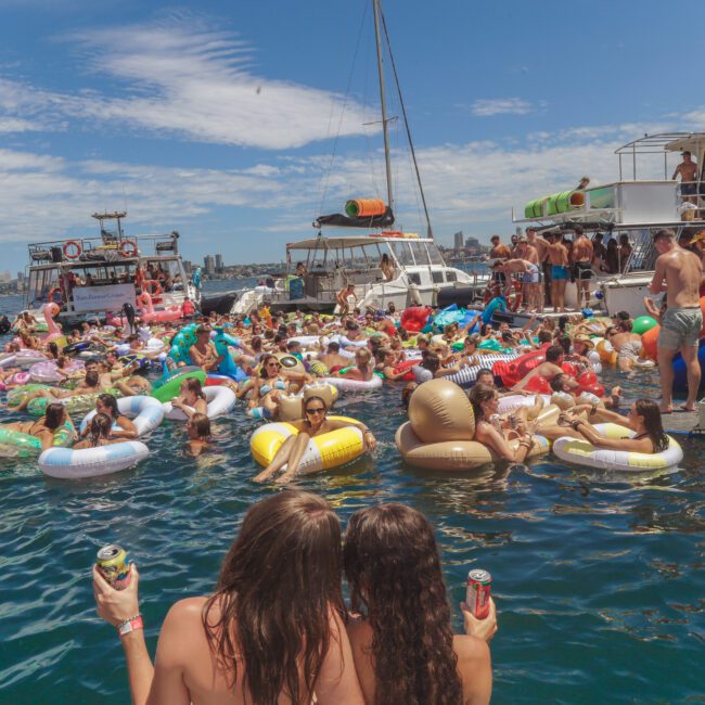 A large group of people relax on colorful inflatable floats in the water near docked boats on a sunny day. Two women holding drinks face the crowd, while others enjoy the party atmosphere. "Yacht Social Club" logo is visible.