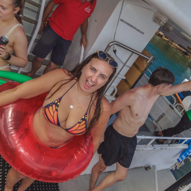A smiling woman in a colorful bikini and red swim ring stands on a boat deck, with other people around her. Sunglasses rest on her head, and the background shows water and part of the boat railing.