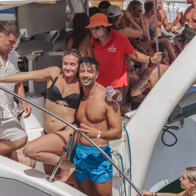 A group of people in swimsuits are smiling and posing on a crowded boat in sunny weather, enjoying a lively social gathering or party on the water.