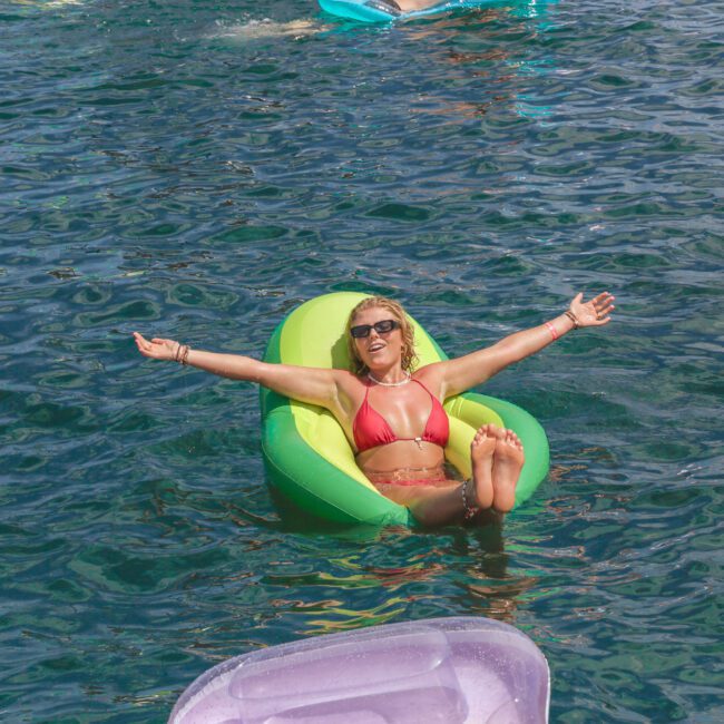 A woman in a red bikini relaxes on a green and yellow inflatable float in clear blue water, arms outstretched and smiling. Other people float nearby on inflatables under the sun.