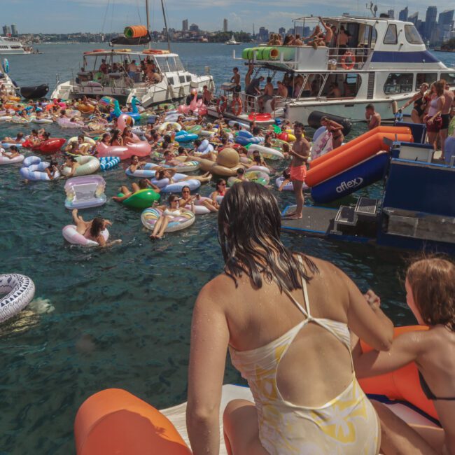 People enjoying a lively pool party in the water between docked yachts, many relaxing on colorful inflatables, with others socializing on the boats under a sunny sky.