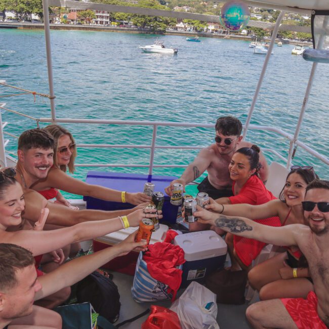 A group of smiling people in swimwear sit on a boat, raising drinks in a cheerful toast. The boat is on clear blue water with a scenic shoreline and yachts in the background.